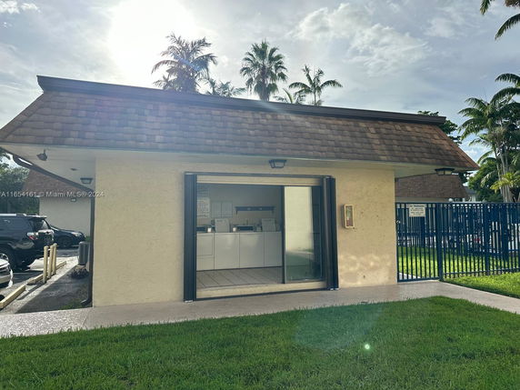 Front view of a small building with an open door, located next to a parking area and surrounded by a fence.