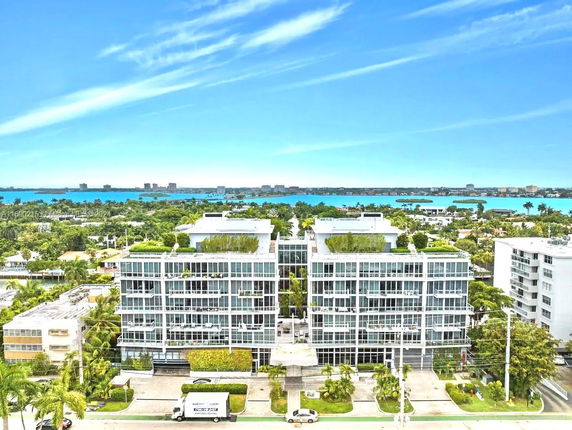 Front view of a modern multi-story building with glass facade and greenery, with water and other buildings in the background.