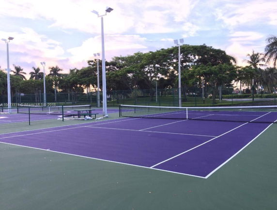 Tennis courts with purple surfaces and surrounding trees under a cloudy sky.