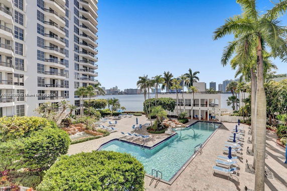 View of a swimming pool area next to a high-rise building with lounge chairs and palm trees.