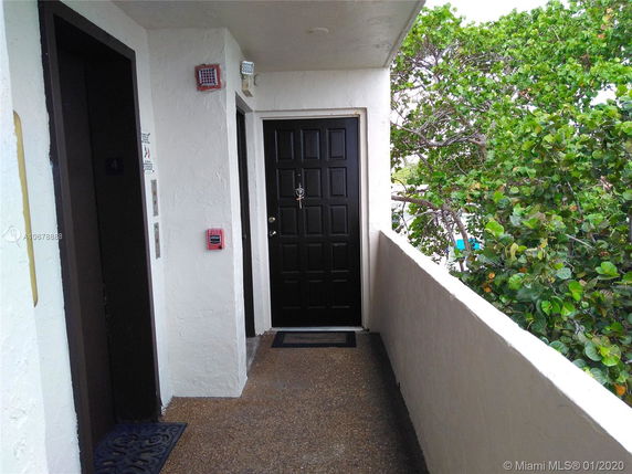 Exterior corridor with an elevator and a black door, overlooking green foliage.
