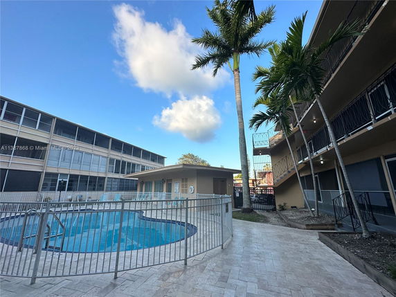 Outdoor swimming pool area with surrounding buildings and palm trees.