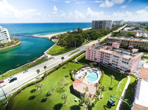 Aerial view of a pink multi-story building with a courtyard pool, set near a coastal waterway and surrounded by green lawns.