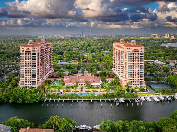 Wide-angle view of two tall apartment buildings with a central building, surrounded by greenery and a waterfront.