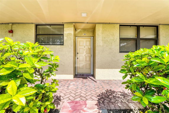 Front view of a building entrance with a beige door and two windows, surrounded by green foliage.
