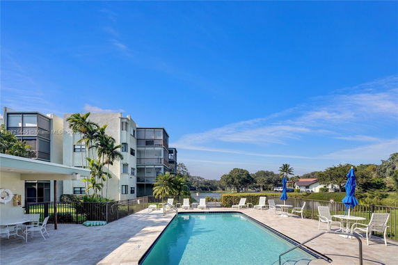 Outdoor swimming pool area adjacent to residential buildings with seating and umbrellas.