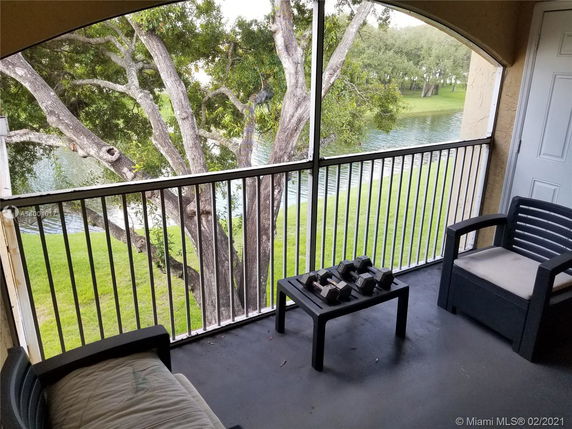 View from a balcony showing a railing, outdoor furniture, and a tree with a body of water and grass in the background.