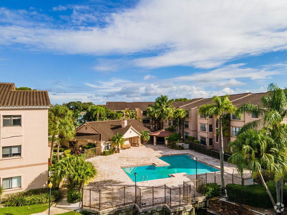 View of a swimming pool area surrounded by multi-story residential buildings with tiled roofs and palm trees.