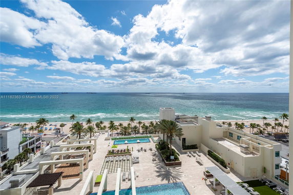Panoramic view of a beachfront area with a building, swimming pools, and palm trees.