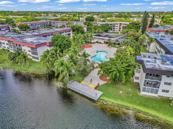 Aerial view of a residential complex with buildings surrounding a central swimming pool and adjacent to a large body of water.