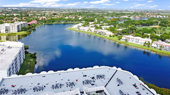 Wide view of a lake with buildings and a complex in the foreground, surrounded by greenery.