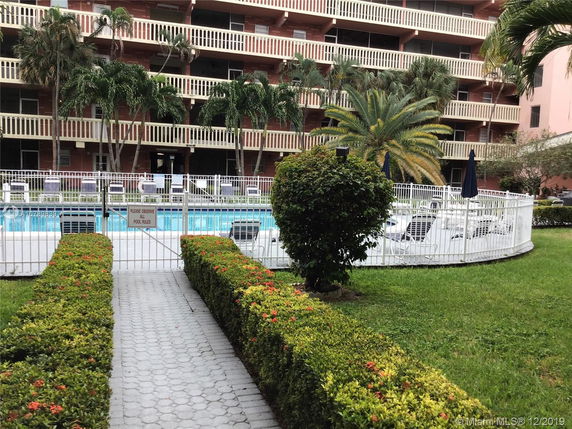 View of a multi-story building with a white fence surrounding a pool area featuring lounge chairs and tropical plants.