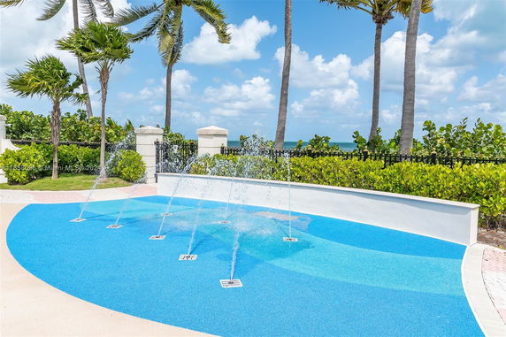 Outdoor play area with water fountains on a blue surface, surrounded by palm trees and greenery.