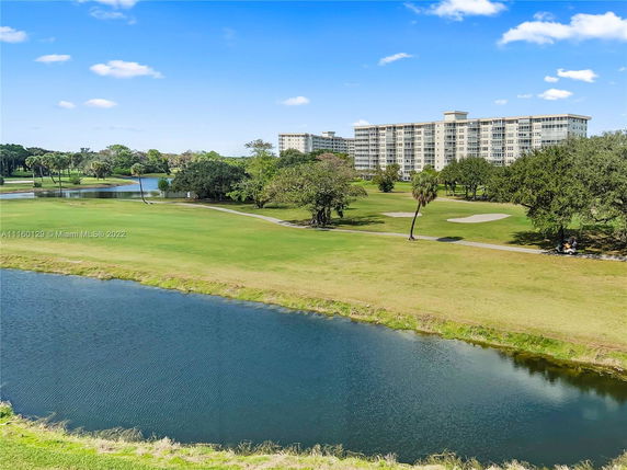 A view of a large residential building surrounded by expansive green fields and a water body in the foreground.