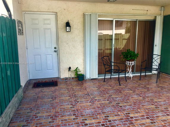 Front view of a house with a white door and sliding glass door, featuring a small patio area with chairs and a plant stand.