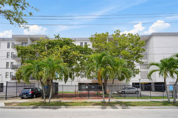 Front view of a multi-story apartment building with balconies, surrounded by trees and a metal fence.