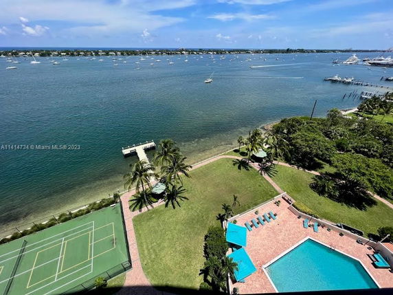 Aerial view of a waterfront area with a swimming pool, tennis courts, palm trees, and a pier extending into the water.