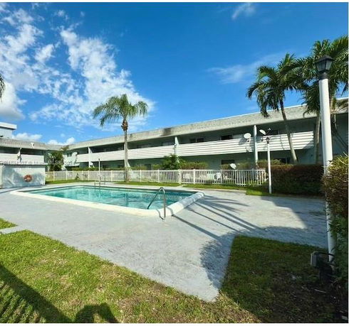 Outdoor view of a swimming pool surrounded by a fence, with a two-story building in the background.