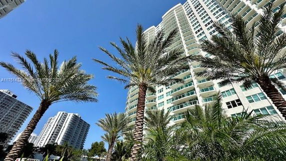 Upward view of a tall modern building with balconies and palm trees in the foreground.