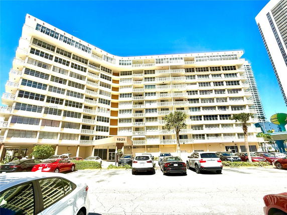 Front view of a multi-story residential building with numerous balconies and a parking area in front.