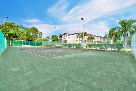Tennis court with fencing and lighting surrounded by trees and a residential building in the background.