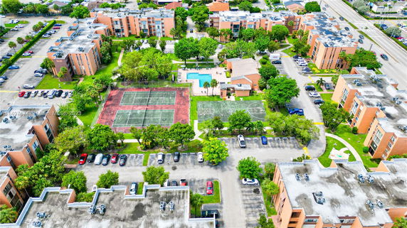 Aerial view of a residential complex with multiple buildings, a swimming pool, tennis courts, parking areas, and surrounding greenery.