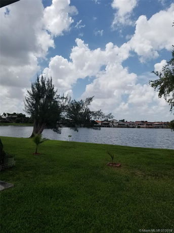 Wide-angle view of a grassy area with a lake and houses in the distance under a cloudy sky.