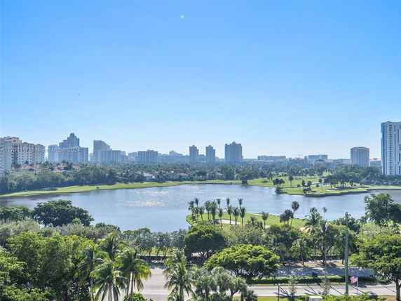 View of a city skyline with high-rise buildings, a lake, and greenery in the foreground under a clear blue sky.