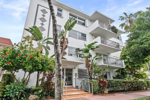 Front view of a multi-story building with balconies and tropical plants in front.