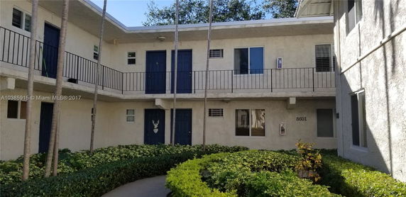 Front view of a two-story building with balconies and blue doors, surrounded by greenery.