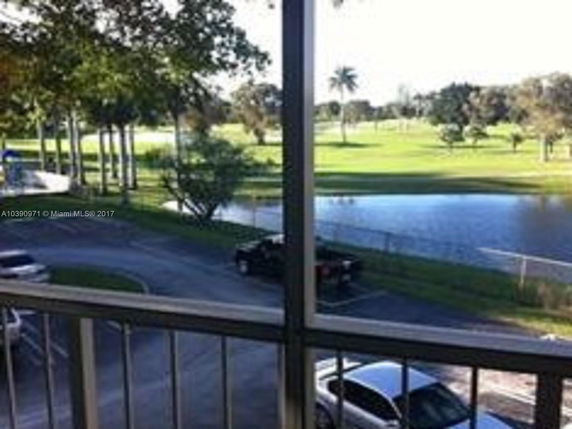 View of a parking lot and grassy area with trees and a pond in the background.
