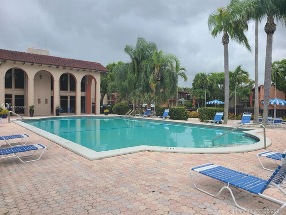 Outdoor swimming pool area with surrounding lounge chairs and nearby building with arched windows.