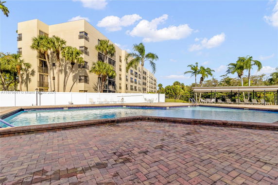 Outdoor swimming pool area with a multi-story building in the background and palm trees.