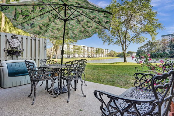 Outdoor seating area with metal chairs and a table under a large umbrella, overlooking a grassy area with trees and a pond. Apartment buildings are visible in the background.