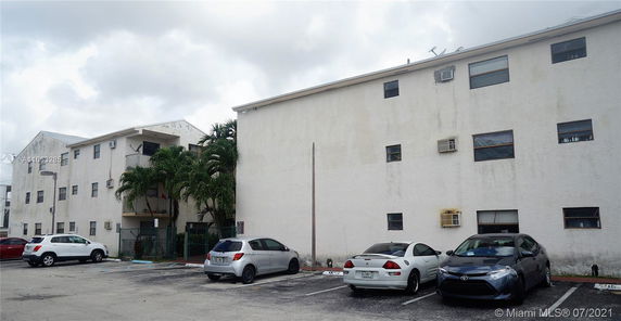 Outdoor parking area with several parked cars and adjacent three-story residential building.