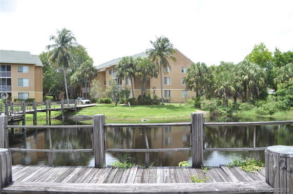 View of a residential building near a water body, surrounded by palm trees and greenery.