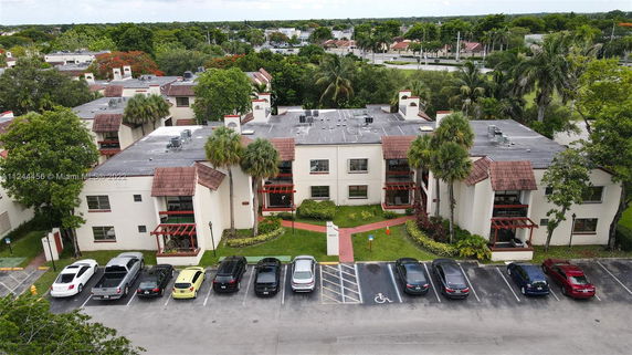 Aerial view of apartment buildings surrounded by trees and greenery, with a parking area in the foreground.