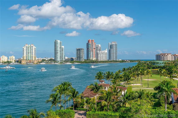 View of a cityscape with tall buildings across a body of water, with boats and palm trees in the foreground.