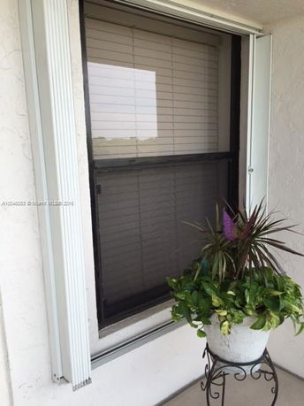 Exterior view of a window with closed blinds and a potted plant on a stand beside it.