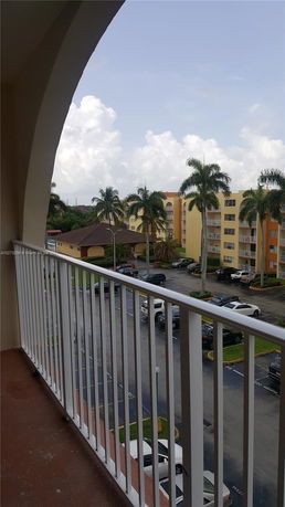 View from a balcony overlooking a parking area and multiple apartment buildings with palm trees nearby.
