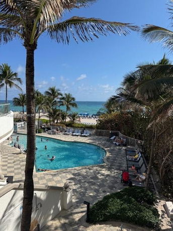 Outdoor swimming pool area with people swimming, surrounded by palm trees and a view of the ocean.