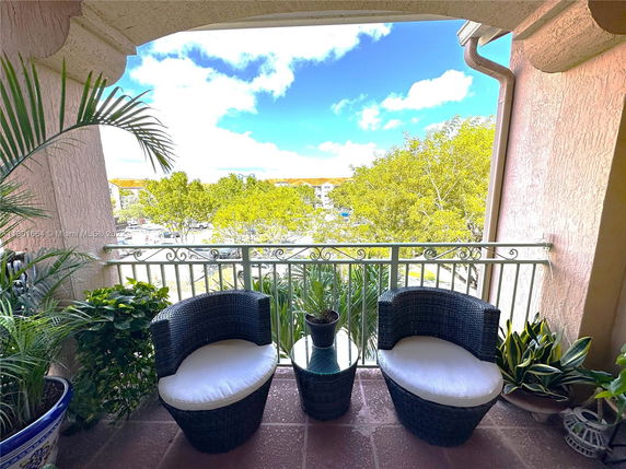 A balcony with outdoor seating overlooking green trees and a parking area.