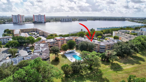 Aerial view of a residential complex near a water body with multiple buildings, a swimming pool, and surrounding greenery.