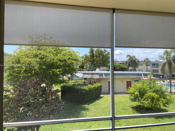View from a window with roller blinds partially down, overlooking a courtyard with trees, a small building, and a swimming pool in the distance.