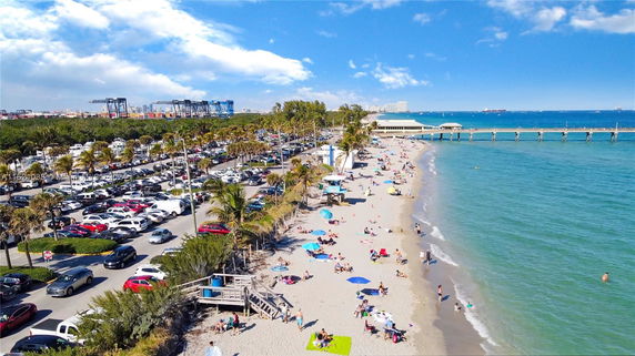 Aerial view of a beach with a parking lot and pier on a sunny day.
