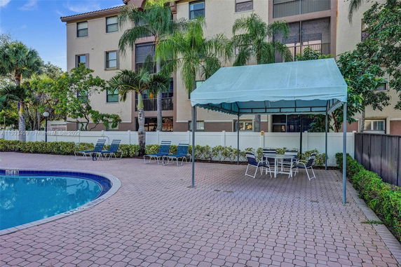 Swimming pool area with patio furniture and green canopy near a multi-story building.