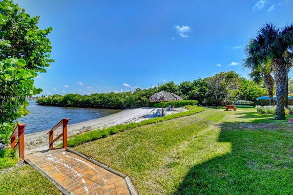 View of a grassy area leading to a sandy beach with a thatched umbrella beside the water.