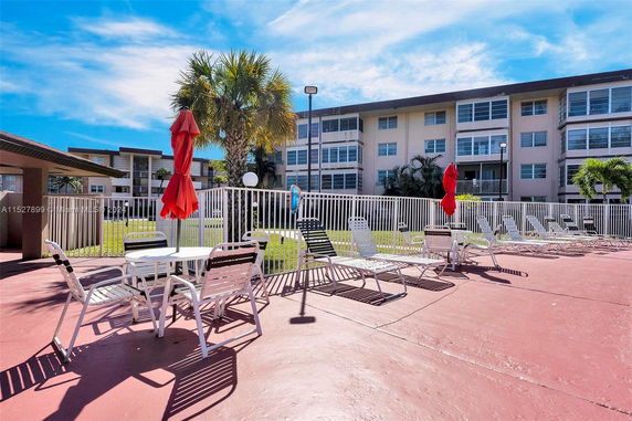 Outdoor area with lounge chairs and patio furniture near a fenced swimming pool, adjacent to a multi-story building.