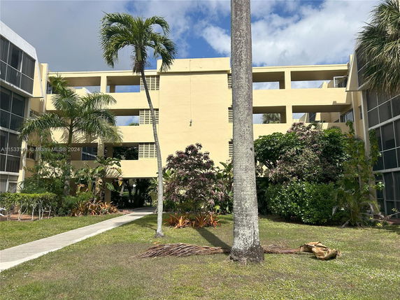 Front view of a multi-story apartment building with open balconies and surrounding greenery.