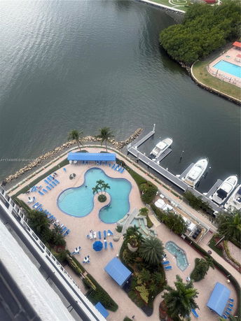 Aerial view of a swimming pool area with blue lounge chairs and palm trees near a waterfront with docked boats.